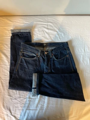 Mörkblå selvedge jeans - Snygga mörkblå jeans från Neuw med klassisk femficksmodell och kontrastsömmar. Jeansen har upprullade benslut och är tillverkade i ett robust denimtyg. Perfekta för dig som gillar stilrena och tidlösa jeans.