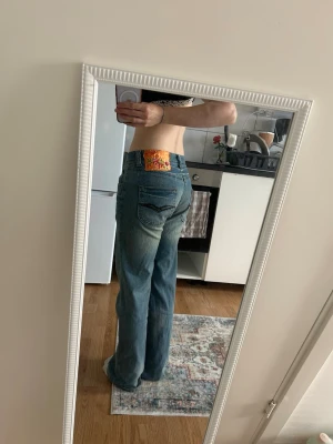 Blå bootcut jeans med färgglad patch - Säljer ett par blå bootcut jeans med låg midja och snygg tvätt. Jeansen har en färgglad patch bak i midjan och klassiska fickor både fram och bak. Perfekta för dig som gillar en avslappnad och trendig look.