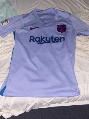 Ljuslila FC Barcelona matchtröja Nike - Säljer en ljuslila FC Barcelona matchtröja från Nike med Dri-Fit-material. Tröjan har klubbmärke, Nike-logga och Rakuten-tryck på bröstet samt La Liga-märke på ärmen. Perfekt för fotbollsträning eller att bära som supporter.