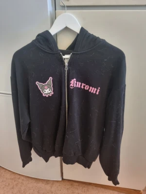 Svart Kuromi hoodie med dragkedja - Svart hoodie med dragkedja och huva, prydd med Kuromi-tryck fram och stort Kuromi-motiv samt text på ryggen i rosa. Mjuk bomullskänsla och ribbade muddar. Perfekt för dig som älskar kawaii-stil och Sanrio-karaktärer.
