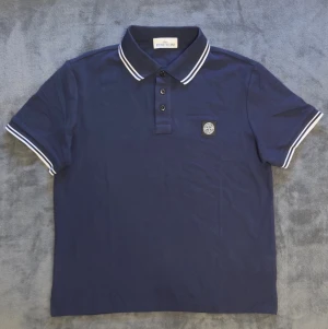 Stone Island pikétröja  - Aldrig använd, Snygg marinblå polotopp från Stone Island med klassisk krage och tre knappar framtill. Toppens ärmar och krage har vita ränder som ger en sportig look. Materialet är mjukt och känns som bomull. Liten Stone Island-logga på bröstet. Pris kan diskuteras