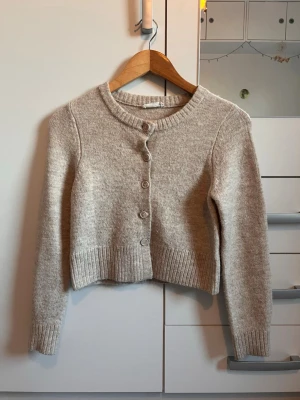 Beige kofta från MNG med knappar - Mysig beige kofta från MNG med rund halsringning och knäppning framtill. Koftan är kortare i modellen och har ribbstickade muddar vid ärmslut och nederkant. Perfekt att slänga över en topp för en enkel och stilren look.