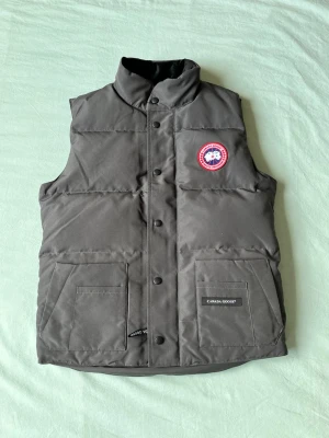 Mörkgrå dunväst från Canada Goose - Snygg mörkgrå dunväst från Canada Goose med hög krage, två stora framfickor och klassisk logotyp på bröstet. Västen har tryckknappar framtill och är tillverkad i slitstarkt material med dunfyllning för extra värme.