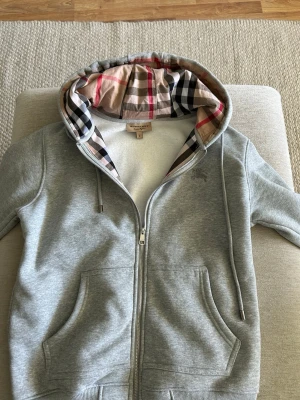 Grå hoodie från Burberry med dragkedja - Snygg grå hoodie från Burberry med klassiskt rutigt foder i huvan och dragkedja framtill. Hoodien har två fickor, ribbade muddar och justerbar dragsko i huvan. Perfekt för en stilren och trendig look.