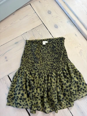 Grön leopardmönstrad kjol H&M - Kort kjol från H&M i grönt med svart leopardmönster. Kjolen har smockad midja och volang nedtill för en cool och trendig look. Perfekt för dig som gillar djurmönster och vill sticka ut lite extra.