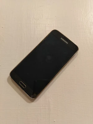 Samsung Galaxy i guld - Snygg Samsung Galaxy med guldfärgad baksida och svart framsida. Telefonen har en klassisk rektangulär form, stor skärm och en bakre kamera med blixt. Baksidan är mönstrad och tillverkad i plast. Perfekt för dig som gillar stilren design och Android.
