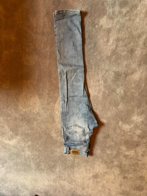 Ljusblå jeans från Replay - Säljer ett par ljusblå jeans från Replay med klassisk femficksdesign och snygga slitningar. Jeansen har en bakficka med broderad detalj och läderpatch med Replay-logga vid midjan. Perfekta för en avslappnad och trendig stil. Ny pris 700kr.
