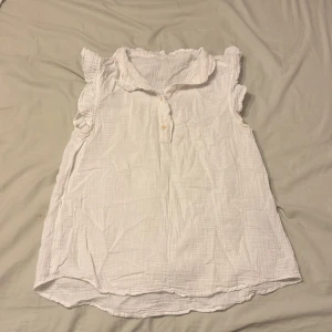 Vit blus med volangärmar och krage - Supersöt vit blus med krage och kort knappslå framtill. Blusen har volangdetaljer vid ärmöppningarna och är ärmlös. Materialet är mjukt och lätt, troligen bomull, med en lite skrynklig struktur. Perfekt till jeans eller kjol för en chill och fräsch look.