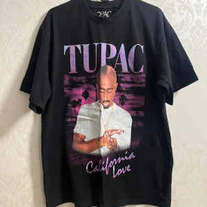 Svart Tupac California Love t-shirt - Svart t-shirt med stort tryck av Tupac och texten 'California Love' i lila och vitt. Klassisk passform med rund hals och korta ärmar. Materialet är mjuk bomull och motivet har en cool retro-vibe med palmer i bakgrunden.