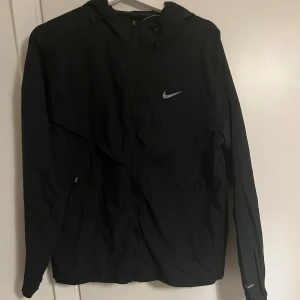 Svart Nike vindjacka med huva - Svart vindjacka från Nike med huva och dragkedja framtill. Jackan har en stilren look med Nike-logga på bröstet, praktiska sidofickor med dragkedja och justerbar huva. Perfekt för dig som gillar sportig och clean stil.
