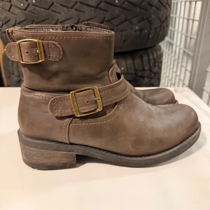Bruna boots med spännen och dragkedja - Snygga bruna boots i skinn med rund tå, låg klack och två dekorativa mässingsfärgade spännen. Dragkedja på insidan för enkel påtagning. Perfekta till jeans eller kjol för en cool vardagslook.