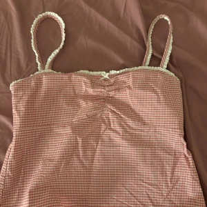 Rutig rosa linnetopp med spetsdetaljer - Supersöt linnetopp i rött och vitt rutigt mönster med smala axelband och spetskant längs halsringningen. Toppen har en liten rosett framtill och rynkade detaljer vid bysten. Perfekt för sommarens alla häng och chill.