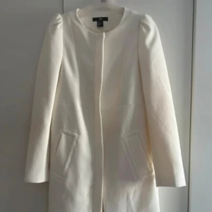 Vit kappa från H&M med dragkedja - Snygg vit kappa från H&M med rund halsringning och diskret dragkedja framtill. Jackan har puffade axlar, raka långa ärmar, två fickor fram och är helfodrad. Materialet är mjukt och känns lyckligt. Perfekt för dig som vill ha en clean och modern look. Tyvärr har kappan tre små defekter