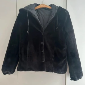 Vändbar täckjacka med fickor  - Svart täckjacka med mjuk fuskpäls på ena sidan och quiltad på andra sidan. Jackan har huva med snörning, tryckknappar framtill och långa ärmar. Perfekt för dig som gillar en chill och varm look under höst och vinter