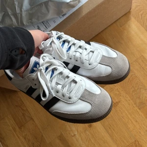 Adidas Samba vita och svarta sneakers - Adidas Samba sneakers i vitt skinn med svarta ränder och grå mockadetaljer på tån. Säljes då de inte kommer till användning, använda 1 gång. Nypris 1349kr