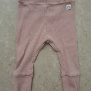 Byxor - Supersöta ribbade babybyxor från Lindex i en mjuk, ljusrosa nyans. Byxorna har breda muddar vid bensluten och en liten tygpatch med regnbågsmotiv framtill. Tillverkade i bomull med lite elastan för extra stretch och komfort.