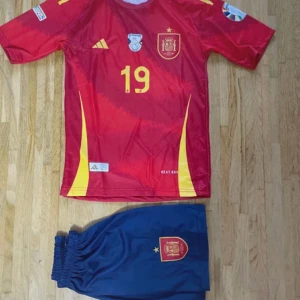 Spanien Lamine Yamal fotbollströja set - Säljer ett officiellt Spanien fotbollströja set med Lamine Yamal och nummer 19. Tröjan är röd med gula detaljer, korta ärmar och tryckta emblem samt Adidas-logga. Matchande marinblå shorts med Spanien-märke ingår. Materialet är lätt polyester, perfekt för match eller träning.