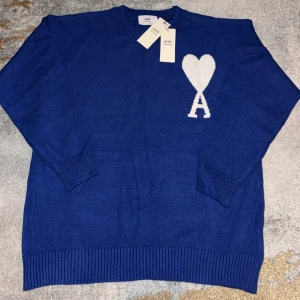 Blå stickad tröja från Ami Paris - Snygg blå stickad tröja från Ami Paris med vit hjärtlogga och bokstaven A på bröstet. Tröjan har rund halsringning, ribbade muddar och är långärmad. Perfekt för dig som vill ha en stilren men ändå unik look.