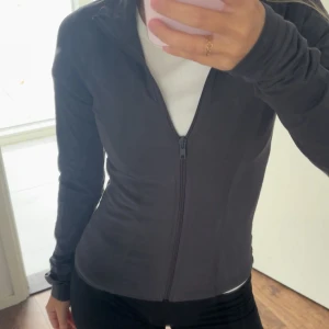 Mörkgrå träningsjacka med dragkedja - Säljer en mörkgrå, slimfit träningsjacka med hel dragkedja framtill. Jackan har lång ärm och står snyggt mot kroppen, perfekt för dig som gillar sportig och clean stil. Materialet är mjukt och stretchigt, vilket gör den skön att bära under aktiva dagar. Ginas soft touch kollektion!