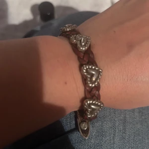 Brunt flätat armband med hjärtan - Unikt brunt flätat armband med silverfärgade hjärtan och detaljer. Armbandet har en cool hästskoformad låsning och flera dekorativa metalldetaljer längs bandet. Perfekt accessoar för dig som gillar westernstil och personliga smycken.