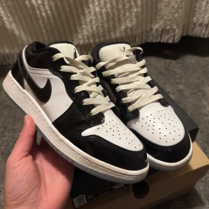 Nike Air Jordan 1 Low svart/vit - Snygga Nike Air Jordan 1 Low i svart och vitt med klassisk design. Skorna har en låg siluett, snörning och perforerad tåbox. Ovandelen är i skinn med blanka svarta paneler och vit bas. Ikonisk Swoosh på sidan och Jumpman-logga på plösen.