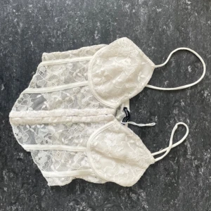 Vit spetskorsett från Bershka - Säljer en vit korsett från Bershka i storlek XS. Toppen har tunna axelband, genomskinlig spets med blommigt mönster och markerade sömmar framtill. Den har en snygg v-ringad form och är perfekt för att skapa en trendig look.