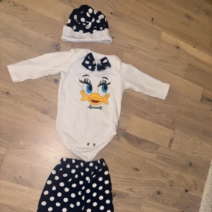 Daisy Duck Set - Body, Byxor & Mössa ankset - Ett sött set med en vit body med Daisy Duck tryck och en matchande rosett. Setet inkluderar även ett par mörkblå byxor med vita prickar och en matchande mössa. Säljer ett par supersöta mjukisbyxor från Miniworld i mörkblått med stora vita prickar. Byxorna har resår i midjan och är gjorda i mjuk bomull, perfekta för bebisar som vill vara både bekväma och stiliga.