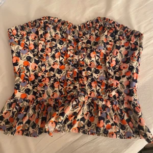 Blommig korsett från H&M med volang - Säljer en blommig korsett från H&M i storlek 36. Toppen har volangkant nedtill och längs överdelen, knappar framtill och ett färgglatt mönster. Perfekt för dig som vill sticka ut med en feminin och trendig look.