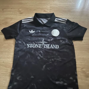 Svart fotbollströja Stone Island x Adidas - Snygg svart fotbollströja från Adidas i samarbete med Stone Island. Tröjan har silvriga detaljer, klassiska Adidas-ränder på axlarna och ett unikt mönster över hela tyget. Broderad logga på bröstet och polokrage. Tillverkad i lätt och ventilerande material.