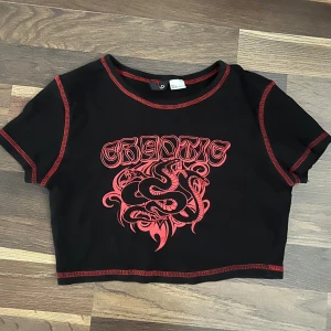 Svart croppad topp med röd drakprint - Cool svart croppad topp från H&M Divided med röd kontrastsöm och ett fett drakmotiv samt texten 'CHAOTIC' framtill. T-shirten har korta ärmar och är gjord i mjuk bomull. Perfekt för dig som gillar edgy streetstyle och vill sticka ut.