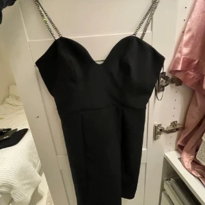 Svart mini klänning med strassband - Supersnygg svart kort klänning med figurnära passform och glittriga strassband som axelband. Klänningen har en hjärtformad urringning och dragkedja i ryggen. Perfekt för dig som vill sticka ut med en elegant och trendig look.