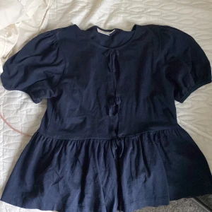Mörkblå blus med knyt från Gina Tricot - Säljer en mörkblå blus från Gina Tricot i storlek 158/164. Blusen har puffiga korta ärmar, rund hals och fina knytband framtill. Den har en volang nedtill som ger en söt och trendig look. Perfekt för dig som gillar stilrena och feminina toppar.