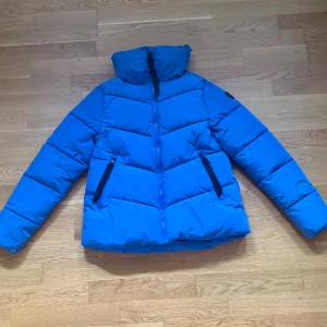 Blå pufferjacka från Everest - Blå pufferjacka från Everest med hög krage och svarta dragkedjor. Jackan har två fickor framtill och en diskret logotyp på ärmen. Materialet är syntet och jackan har en sportig look, perfekt för kyliga dagar.