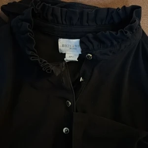 Svart henley med volangkrage Holly & Whyte - Svart långärmad henley från Holly & Whyte med snygg volangkrage och knappar framtill. Skjortan har en clean look och är tillverkad i mjukt material som känns skönt mot huden. Perfekt för dig som gillar stilrena detaljer och vill ha något extra i kragen.