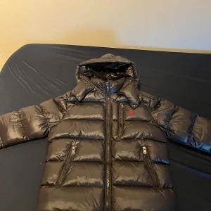 Svart pufferjacka från Polo Ralph Lauren - Snygg svart pufferjacka från Polo Ralph Lauren med huva och dragkedja framtill. Jackan har två sidofickor med dragkedja och ett rött Polo-logomärke på bröstet. Materialet är glansigt och jackan är quiltad för extra värme. Perfekt för kalla dagar. Luvan är avtagningsbar har bara ägt jackan i 2 veckor 