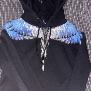 Svart hoodie Marcelo Burlon med blå vingar - Svart hoodie från Marcelo Burlon County of Milan med ikoniskt blått och vitt vingmönster över axlar och hals. Tröjan har huva med snörning och lång ärm. Perfekt för dig som vill sticka ut med en cool streetwear-look.