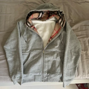 Grå hoodie från Burberry  - Säljer en grå hoodie från Burberry med klassiskt rutigt foder i huvan och längs insidan. Tröjan har broderad Burberry-logga på bröstet, två fickor fram, snörning i huvan och hel dragkedja. Perfekt för dig som gillar stilrena detaljer och ikoniskt mönster. Storlek L men kan också passa M.