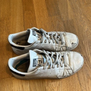 Adidas Stan Smith vita sneakers - Klassiska Adidas Stan Smith sneakers i vitt skinn med mörkblå detalj på hälen och perforerade sidor. Skorna har rund tå, platt sula och snörning framtill. Ikonisk Stan Smith-logga på plösen och hälen. Perfekta för en avslappnad streetstyle.