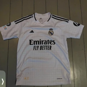 Real Madrid Bellingham matchtröja - Vit Real Madrid fotbollströja från Adidas med svarta detaljer och klubbmärke på bröstet. Tryck med Bellingham och nummer 5 på ryggen. Klassisk V-ringning, korta ärmar och tre svarta ränder på axlarna. Materialet är lätt och andas, perfekt för match eller träning.