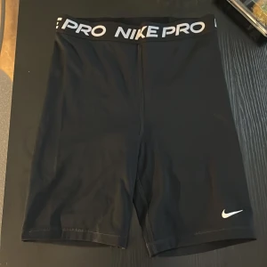 Svarta Nike Pro träningsshorts - Säljer ett par svarta Nike Pro shorts med bred elastisk midja och vit logga. De är tighta och gjorda i ett stretchigt syntetmaterial som passar perfekt för träning. Snyggt och sportigt med Nike Pro-tryck runt midjan.