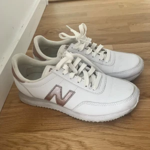 Vita New Balance sneakers med roséguld - Snygga vita New Balance sneakers med detaljer i roséguld på sidorna och hälen. Klassisk modell med snörning, rund tå och platt sula. Perfekt för en clean och sportig look.