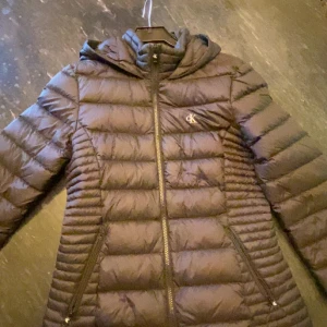 Svart pufferjacka Calvin Klein Jeans - Svart pufferjacka från Calvin Klein Jeans med huva och snygga logodetaljer på bröstet och ärmen. Jackan har två dragkedjeförsedda fickor, quiltad design och en stilren look. Perfekt för dig som vill ha en varm och trendig jacka med cool CK-vibe.