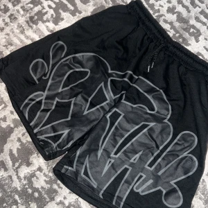 Svarta Syna World shorts 65% polyester 35% bomull - Svarta shorts med stor, grå grafisk print över hela framsidan. De har elastisk midja med grov snörning för extra streetkänsla. Passformen är loose och materialet känns som bomull och polyester,perfekt för chill eller träning. Inga taggar. 