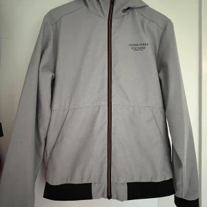 Grå vindjacka från Jack & Jones - Snygg grå vindjacka från Jack & Jones med svart kant nertill och vid ärmslut. Jackan har huva som går att justera, hel dragkedja framtill och vattenavvisande material. Perfekt för dig som gillar stilren och funktionell design.
