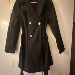 Svart trenchcoat med bälte och knappar - Stilren svart trenchcoat med dubbelknäppning i guldiga knappar och tillhörande bälte i midjan. Jackan har klassisk krage, lång ärm och en figurnära passform som ger en snygg siluett. Perfekt för kyligare dagar under höst och vinter