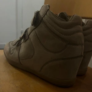 Isabel Marant inspirerade skor - Säljer ett par beige sneakers med kilklack och tre breda kardborreband. Skorna är i mocka och har en sportig men ändå stilren vibe. Perfekta för dig som vill ha lite extra höjd men ändå känna dig bekväm hela dagen.