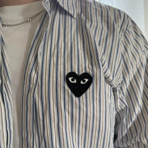 Randig skjorta Comme des Garçons Play - Säljer en vit skjorta med blå och bruna ränder från Comme des Garçons Play. Skjortan har klassisk krage, knappar framtill och det ikoniska svarta hjärta med ögon broderat på bröstet. Tillverkad i mjuk bomull, perfekt för dig som gillar stilrena detaljer.