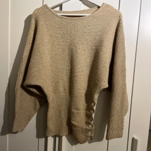 Beige stickad tröja med knappar - Säljer en beige stickad tröja med rund hals och långa ärmar. Tröjan har ribbad midja och dekorativa knappar längs ena sidan nedtill, vilket ger en snygg detalj. Perfekt för lager-på-lager under kyliga dagar.