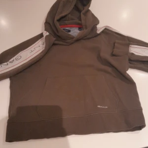 Brun hoodie från GANT med logga - Säljer en brun hoodie från GANT med huva och stor magficka. Snygga vita ränder med GANT-logga längs ärmarna och diskret logotyp framtill. Perfekt för dig som gillar sportig och avslappnad stil.