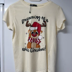 Jultröja  - Krämvit t-shirt från FB Sister med tryck av en söt ren i tomteluva och texten 'Dreaming of a white Christmas'. T-shirten har rund hals, korta ärmar och är gjord i mjuk bomull. Perfekt för dig som vill ha en mysig julstil!